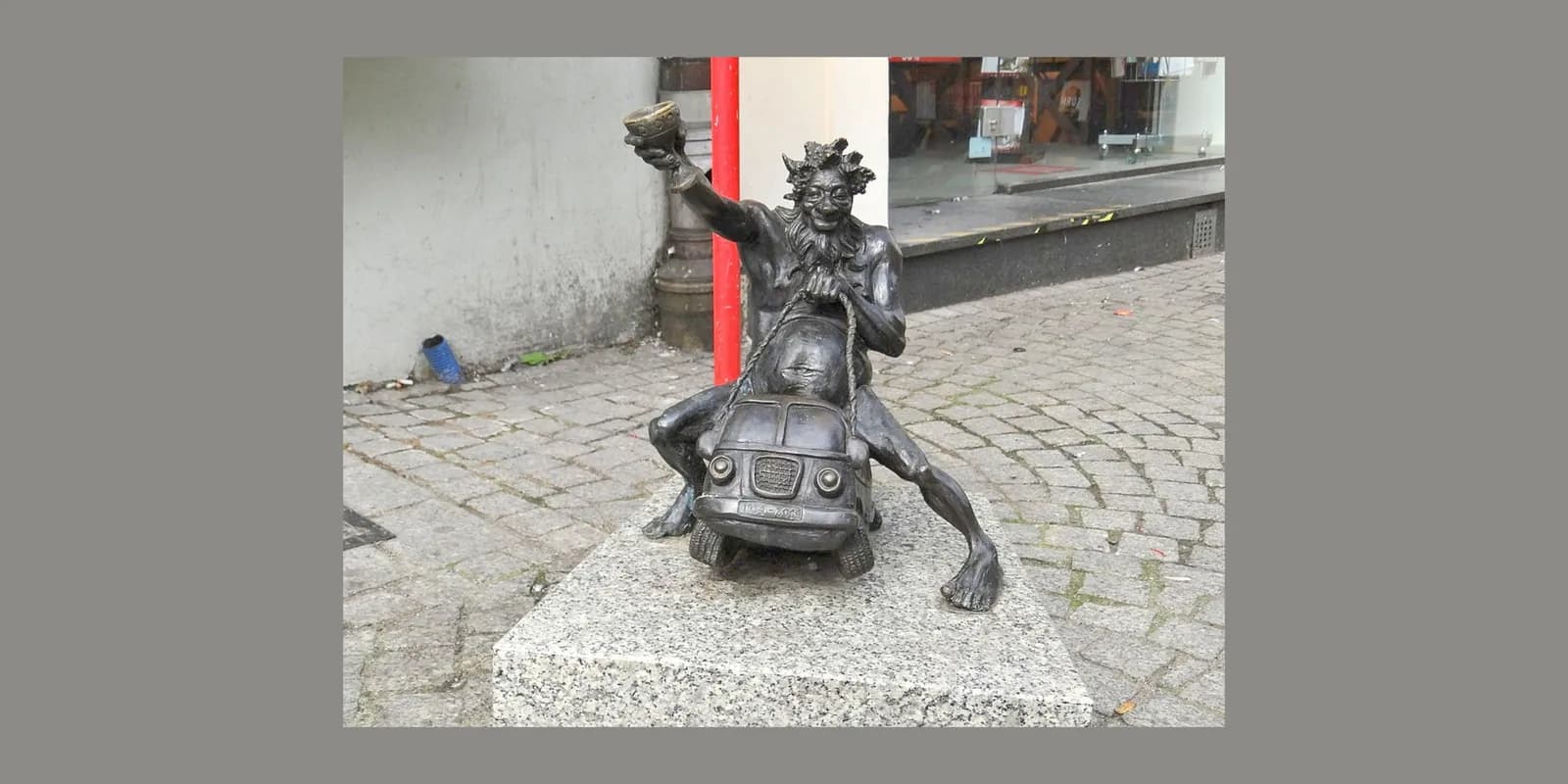 A bronze statue of a satyr-like figure with a crown of leaves, holding up a cup and pulling a small car, stands on a granite base on a cobblestone street.