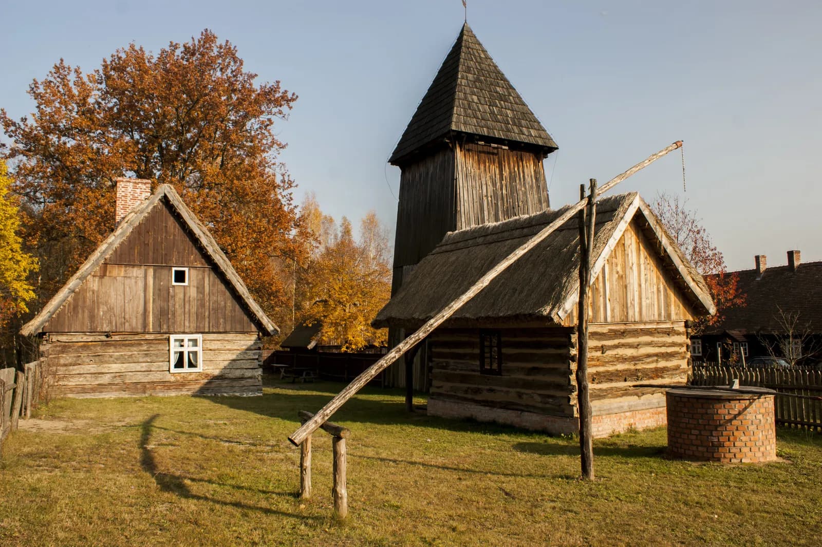 A rural scene featuring a wooden house with a thatched roof, a tall wooden church bell tower, and a well with a wooden shadoof, all set against a backdrop of autumn trees with golden leaves.